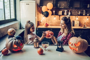 Joyeux halloween! Jolie jeune femme enceinte avec sa petite fille mignonne se préparent à Halloween sur la cuisine. Maman et sa fille s’amusent avec des citrouilles tout en le peignant.