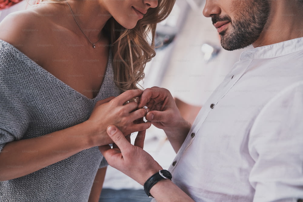 침실에 서 있는 동안 여자 친구의 손가락에 약혼 반지를 끼고 있는 젊은 남자의 클로즈업