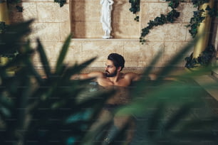 Giovane bell'uomo barbuto che si diverte e si rilassa in un esclusivo resort termale. Si siede nella vasca idromassaggio e beve vino.