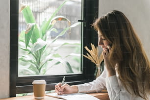 Junge Geschäftsfrau in weißem Kleid sitzt am Tisch im Café und schreibt in Notizbuch. Asiatische Frau spricht Smartphone und Tasse Kaffee. Freiberufler, der im Café arbeitet. Schüler lernen online.