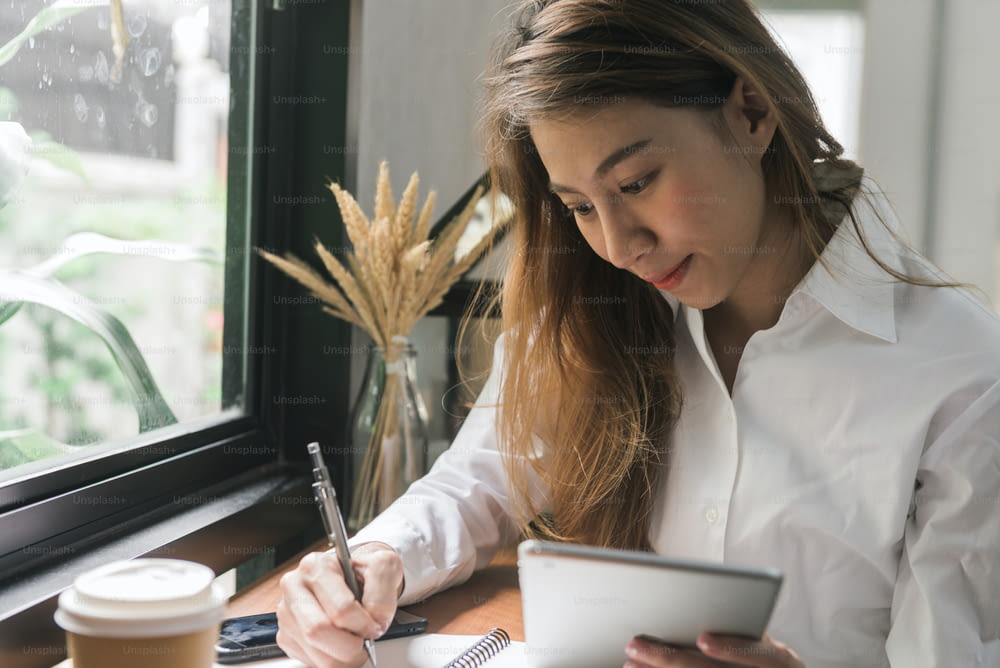 Giovane donna d'affari in abito bianco seduta al tavolo nel caffè e scrivendo nel taccuino. Donna asiatica che usa tablet e tazza di caffè. Libero professionista che lavora in una caffetteria. Apprendimento online degli studenti.
