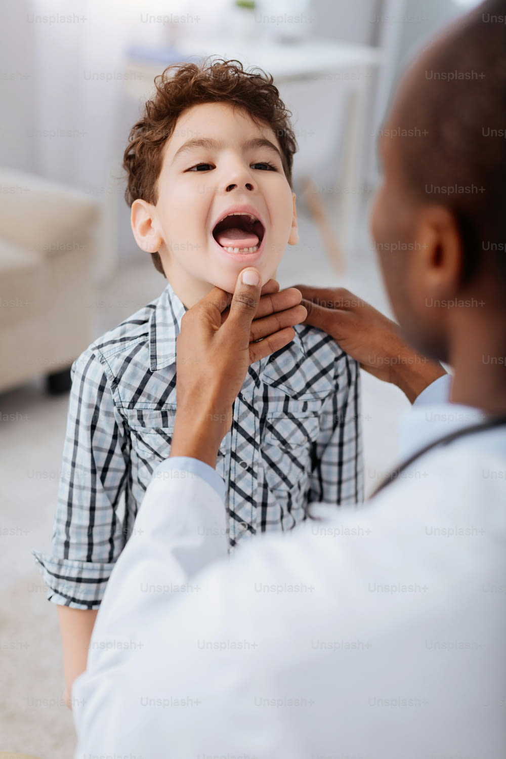 Doctor diagnosis. Boy standing while opening mouth and experienced professional male doctor touching his chin