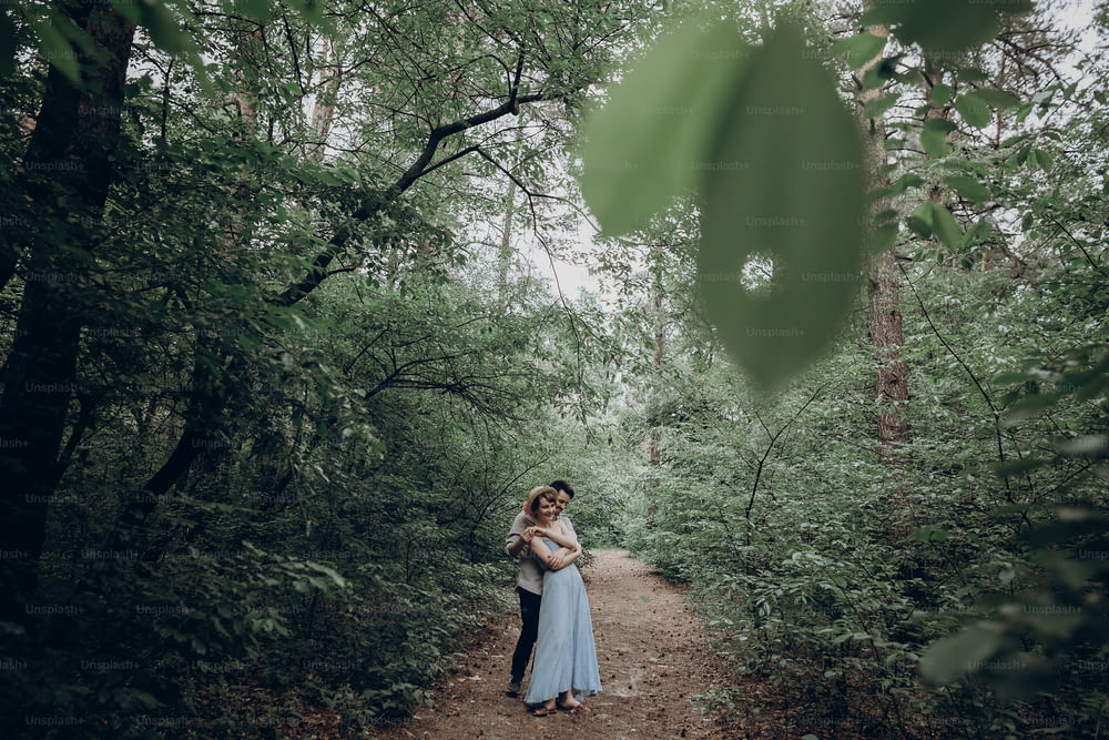 stylish hipster bride and groom walking and dancing in green summer forest. happy couple in love, modern outfit, relaxing at park. girl in dress and straw hat with peony. rustic wedding concept