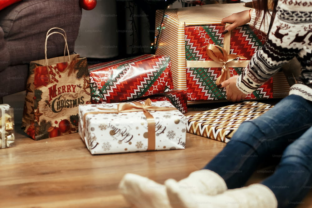 Mujer apilando regalos de Navidad debajo del árbol. ideas para regalos. Chica en suéter con ciervos y con sombrero de Papá Noel en sala festiva con luces. Concepto de saludos de temporada. Momento alegre