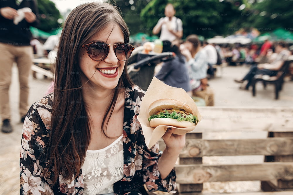 elegante donna hipster in occhiali da sole con labbra rosse che tiene un hamburger succoso. ragazza boho che tiene l'hamburger e sorride al festival del cibo di strada. estate. Viaggi per le vacanze estive