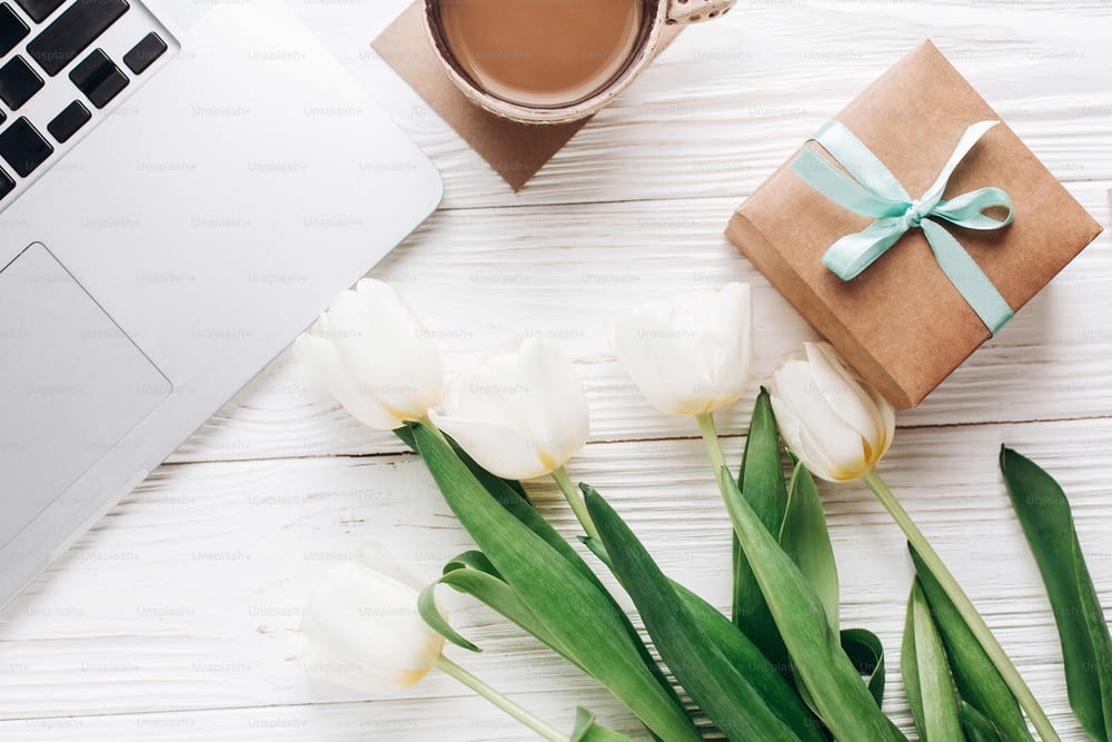 laptop con caffè mattutino e tulipani ed elegante confezione regalo su sfondo rustico in legno bianco. Flat lay freelance, gadget da lavoro con spazio per il testo. vacanze di primavera. Giornata felice della donna