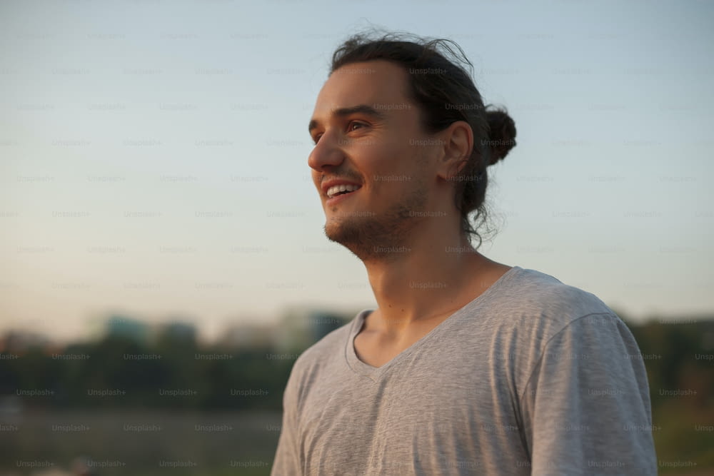 Profilo di bell'uomo sorridente che mostra i denti nel parco, sul lago mentre guarda l'alba. Ritratto di uomo atletico positivo di capoeira sullo sfondo della spiaggia della città, indossando una maglietta grigia.