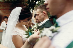 Beautiful bride in vintage wedding dress putting on boutonniere on handsome groom and his stylish groomsmen outdoors before the ceremony, hands and flowers closeup