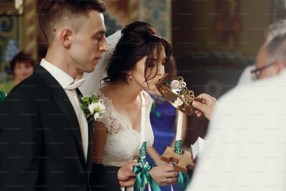 Happy spiritual couple, stylish groom and beautiful brunette bride in white dress holding candles at wedding church ceremony during golden crown coronation