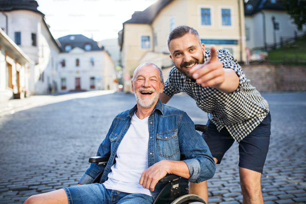 Un fils hipster adulte avec son père âgé en fauteuil roulant lors d’une promenade en ville, pointant du doigt quelque chose.