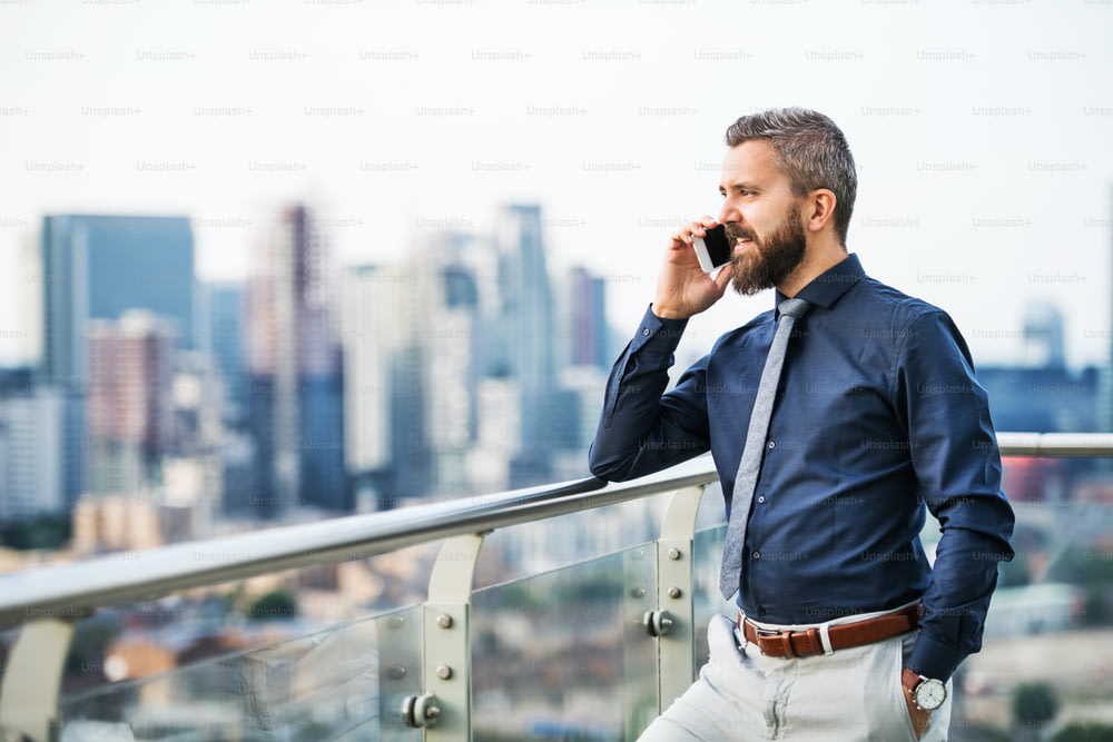 A portrait of businessman with smartphone standing against London rooftop view panorama, making a phone call. Copy space.
