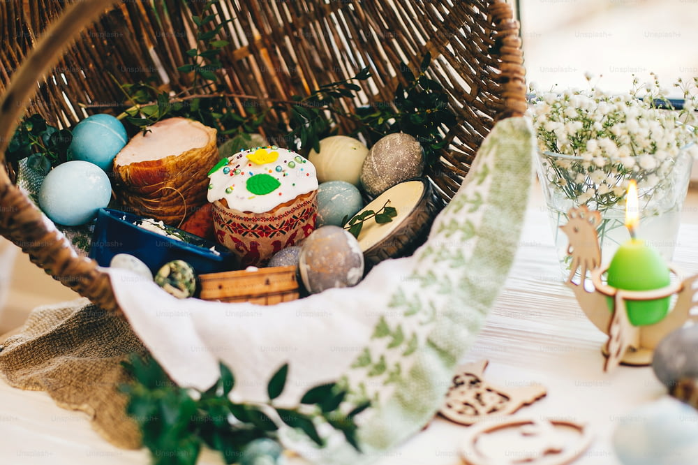 Traditional Easter basket with stylish eggs, easter bread, ham, sausage, butte, and flowers, candle on rustic wooden background. Happy Easter concept. Traditional food in rustic basket