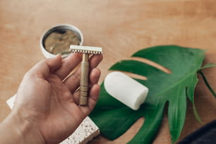 Hand holding reusable vintage razor for shaving on background of natural soap, solid shampoo bar, eco deodorant, sponge on wood with green monstera leaf. Zero waste, plastic free beauty essentials