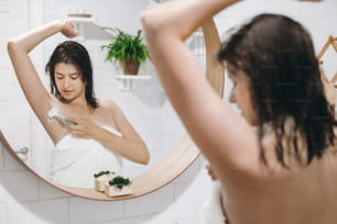 Mulher atraente nova em axilas de barbear toalha branca, olhando no espelho no banheiro elegante. Cuidados com a pele e o corpo. Conceito de Depilação. Mulher após o banho barbear com navalha