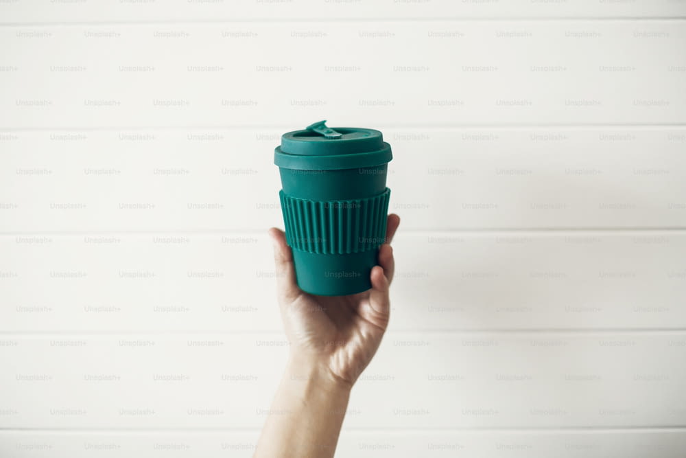 흰색 나무 배경에 세련된 재사용 가능한 에코 커피 컵을 들고 있습니다. 천연 대나무 섬유로 만든 그린 컵, 제로 웨이스트 개념. 일회용 플라스틱을 금지합니다. 선택하십시오. 커피를 가져 가라.
