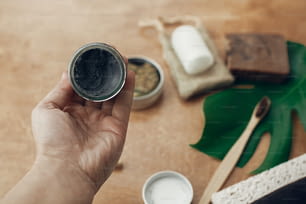 Hand holding natural charcoal toothpaste in glass jar on background of bamboo toothbrush, soap, solid shampoo, metal razor, deodorant on wood with green monstera leaf. Sustainable lifestyle