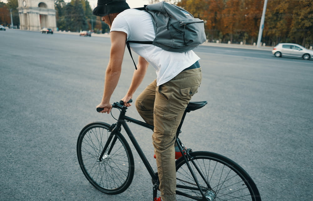 Vista lateral trasera joven moderno, estudiante con mochila, ciclismo en una bicicleta clásica en la carretera de la ciudad. Espacio de copia. Concepto de estilo de vida activo y ecológico