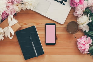 Stilvolles Telefon mit leerem Bildschirm, Laptop, Kaffeetasse, Notizbuch, Geschenk mit rosa und weißen Pfingstrosen auf Tisch flach lag mit Platz für Text. Freiberufliches Konzept. Stilvoller Arbeitsplatz.