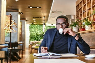 Primo piano di un giovane CEO afroamericano elegante in giacca blu scuro che passa attraverso alcune scartoffie, seduto al tavolo del caffè con documenti e prendendo appunti sul taccuino mentre beve il caffè mattutino nel caffè
