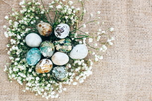 Stilvolle Osterwachteleier mit Frühlingsblumen im Blumennest auf rustikalem Stoff in sonnigem Licht auf Holz. Moderne bunte Eier mit natürlichem Farbstoff in Blau, Grün bemalt. Frohe Ostern, Grußkarte