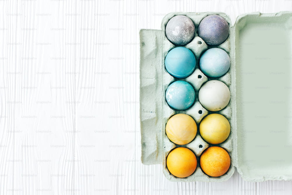 Stylish Easter eggs in carton tray on white wooden background, space for text. Modern colorful easter eggs painted with pastel natural dye in rainbow colors. Happy Easter. Holiday decor