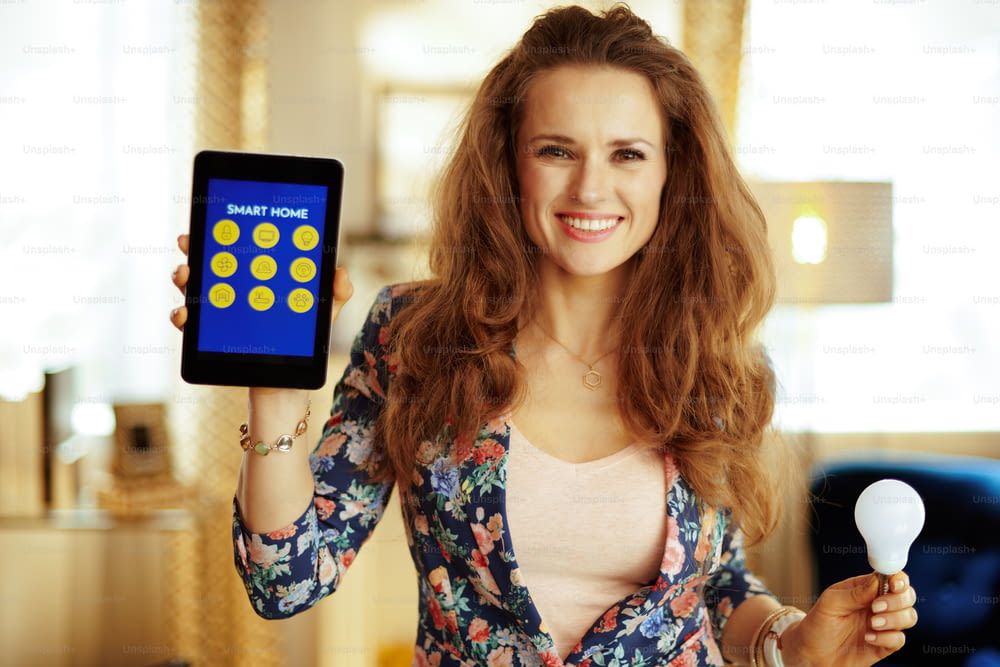 Portrait of smiling young housewife with long brunette hair showing tablet PC with smart home application and smart lamp in the modern living room.