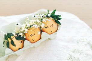 Happy Easter,  eco concept. Stylish Easter eggs with cute faces in floral wreath crowns in carton tray on rustic background. Modern easter eggs with flowers and sleepy eyes in sunny light