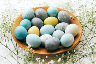 Happy Easter. Stylish Easter eggs with spring flowers in wooden plate on white wooden background. Modern easter eggs painted with natural dye in yellow,blue,green,grey marble colors
