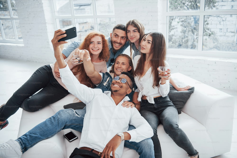 Fare selfie. Giovani amici allegri che si divertono e bevono nell'interno bianco.