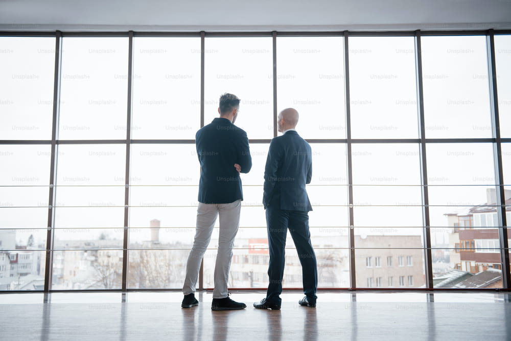 Two businessmen deep in discussion together while standing in an office boardroom with windows overlooking the city.