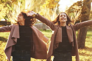 Jóvenes gemelas morenas felices y sonrientes de pie y con las manos abiertas de par en par, su cabello se disipa, vistiendo un abrigo informal en el parque soleado de otoño sobre el fondo borroso claro. Concepto de libertad.