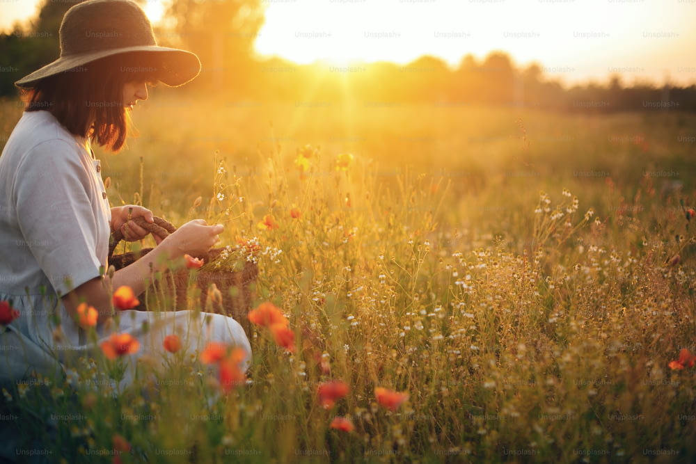 Ragazza elegante in vestito di lino che raccoglie fiori in un cesto di paglia rustico, seduta in un prato di papaveri al tramonto. Donna boho in cappello che si rilassa nella calda luce del sole serale nel campo estivo. Spazio per il testo