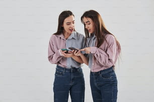 Mostra algumas coisas interessantes em seus telefones. Duas irmãs gêmeas em pé e posando no estúdio com fundo branco.