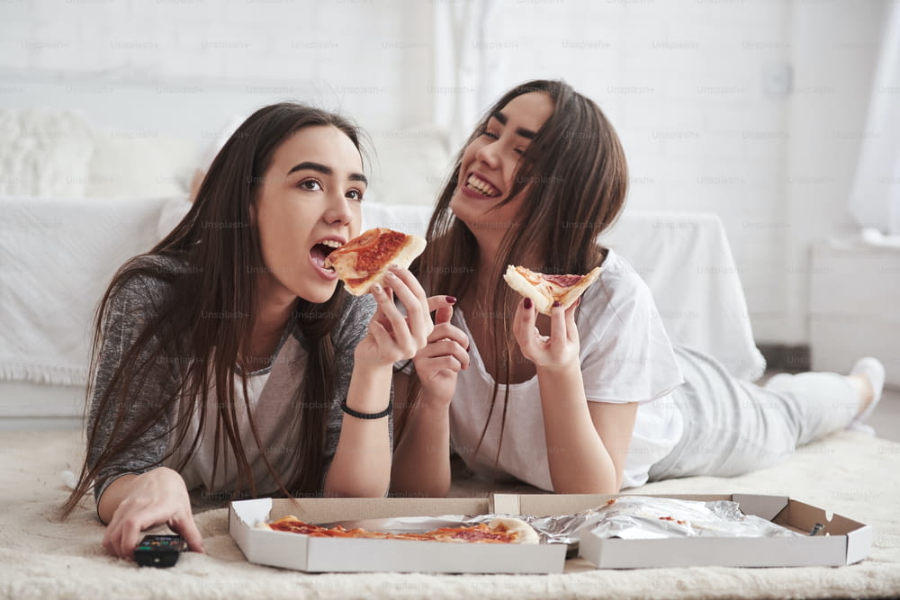 Just having fun. Sisters eating pizza when watching TV while lying on the floor of beautiful bedroom at daytime.