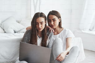 Meninas com aparência séria. Gêmeas jovens do sexo feminino sentadas no chão perto da cama branca e usando laptop de cor prata.
