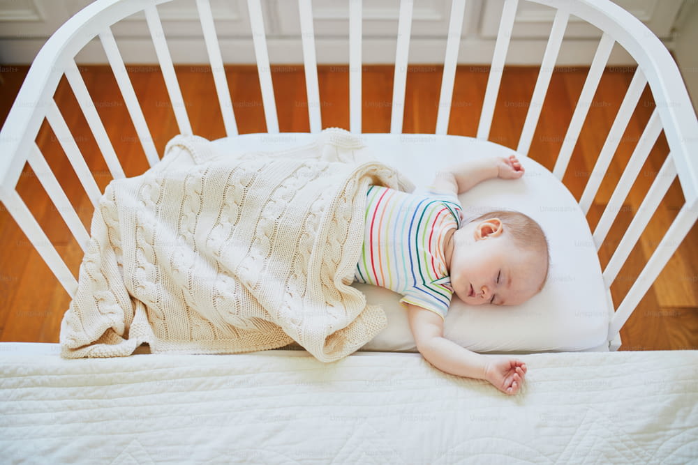 Entzückendes kleines Mädchen, das in einem Co-Sleeper-Kinderbett schläft, das am Bett der Eltern befestigt ist. Kleines Kind mit einem Tagesschlaf im Kinderbett. Schlaftrainingskonzept. Kleinkind im sonnigen Kinderzimmer