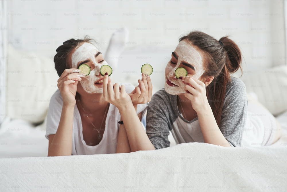 Lying on the whte bed. Conception of skin care by using fresh cucumber rings and white mask on the face. Two female sisters have weekend at bedroom.