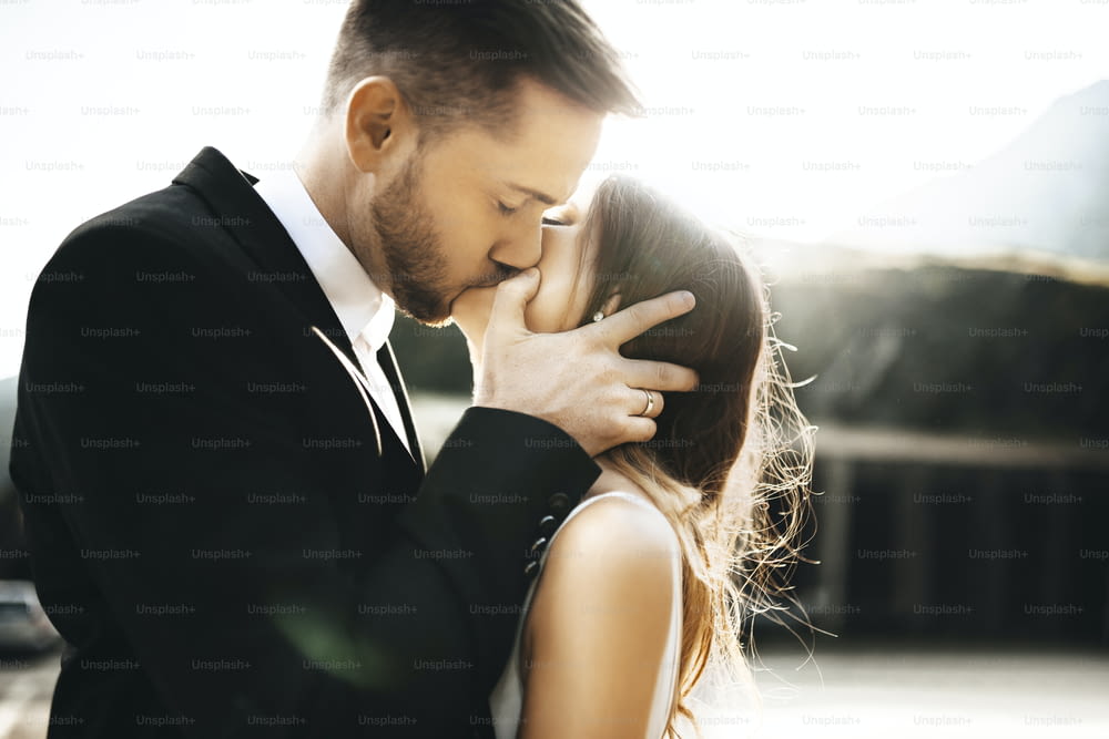 Close up portrait of a handsome young couple kissing in their wedding day against sunrise while groom is holding his bride's face .