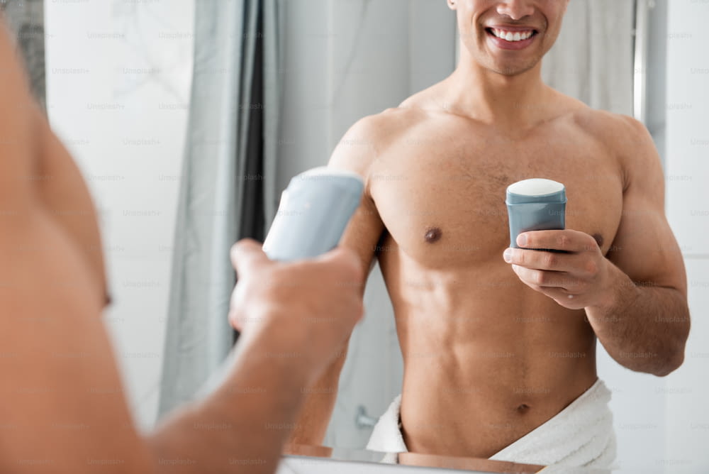 朝の衛生コンセプト。浴室の鏡の前に滞在しながら制汗剤を保持している若い陽気な裸の筋肉質男性の腰の上のトリミングされた頭の反射