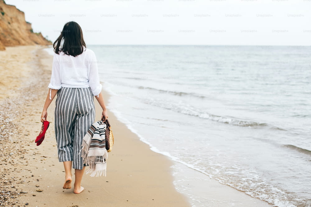 Stylish hipster girl walking barefoot on beach, holding bag and shoes in hand. Happy boho woman relaxing at sea, enjoying walk on tropical island. Summer vacation. Space for text.