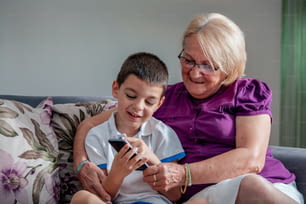 Happy grandmother and cute grandson using cellphone together, smiling older grandma and child girl having fun taking selfie on phone, cheerful granny with little kid play making photo on mobile