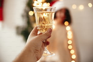 Hombre brindando con copa de champán a una chica feliz en sombrero de Papá Noel con bengala, celebrando en las luces del árbol de Navidad en la sala festiva. Feliz fiesta de fin de año. Felices Fiestas