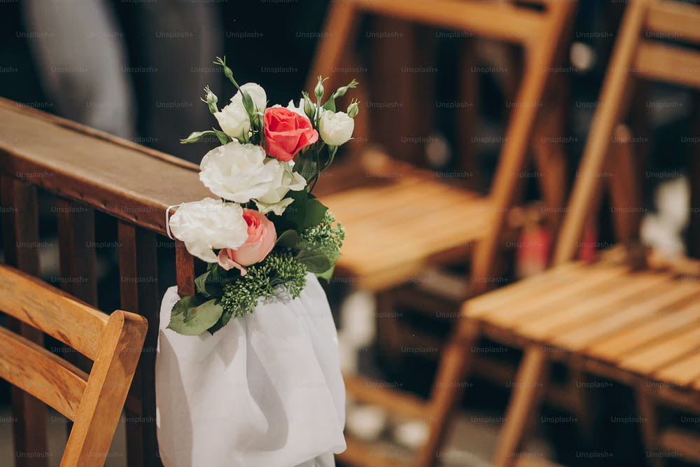 Stilvolle Hochzeitsdekoration von Holzbänken in der Kirche für die heilige Ehe. Schöne Rosen- und Tüllsträuße auf Holzstühlen, Anordnung des Ganges in der Kirche