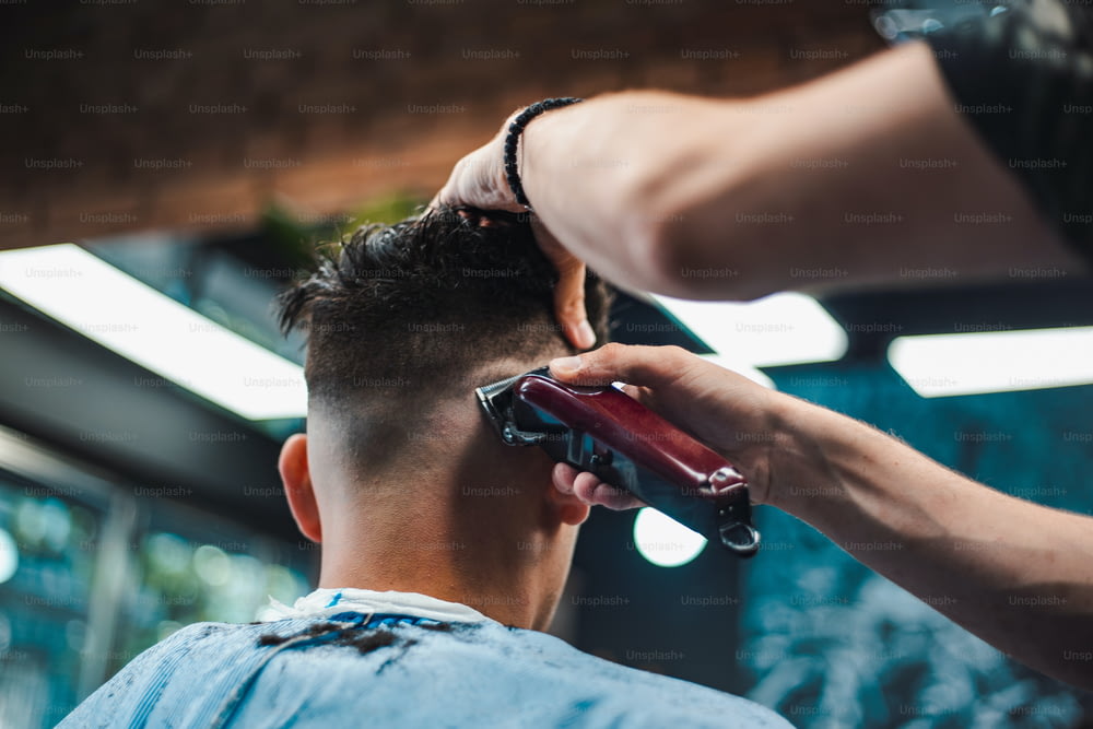 Close up shot of man getting trendy haircut at barber shop. Male hairstylist serving client, making haircut using machine and comb.