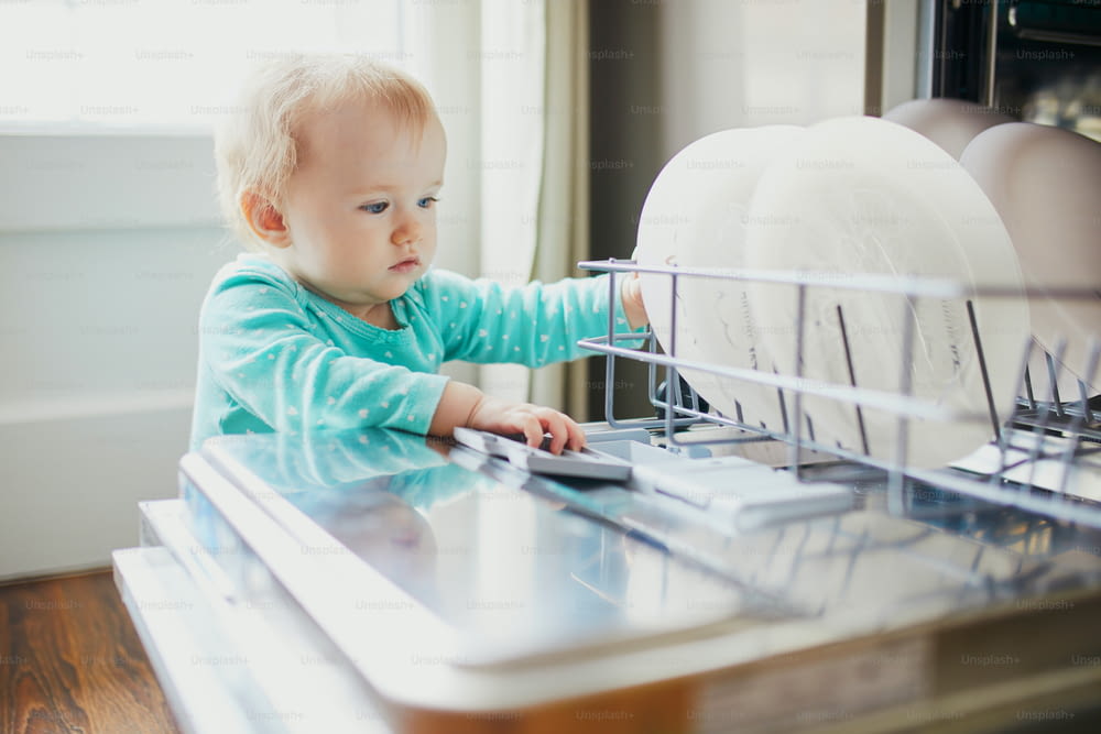 Kleines Kind hilft beim Entladen der Spülmaschine. Kleines Mädchen sitzt auf dem Boden in der Küche. Kleines Kind zu Hause