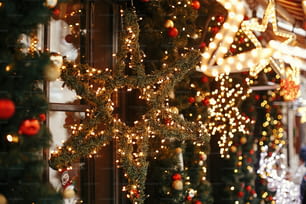 세련된 크리스마스 별이 조명되고 빨간색과 금색 싸구려가 있는 전나무 가지, 도시 거리의 휴가 시장에서 건물 앞에 있는 황금 조명 보케. 크리스마스 거리 장식