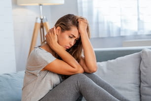 Portrait d’une femme séduisante assise sur un canapé à la maison avec un mal de tête, ressentant de la douleur et avec une expression de malaise.