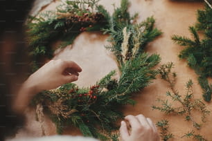 Fazendo grinalda de Natal rústica. Mãos segurando ramos de abeto, e pinhas, linha, bagas na mesa de madeira. Oficina de coroa de Natal. Autêntica natureza morta elegante