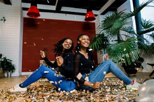 Two african girls, happy stylish friends celebrating new year or birthday party sit back with each other and throw a confetti. Fashion elegance women enjoying time together.