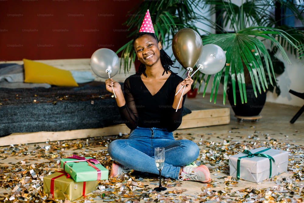 Lustiges afroamerikanisches Mädchen mit Geburtstagshut und Luftballons auf dem Boden sitzend mit Geschenkboxen, Geschenken und Konfetti. Feier, Partykonzept